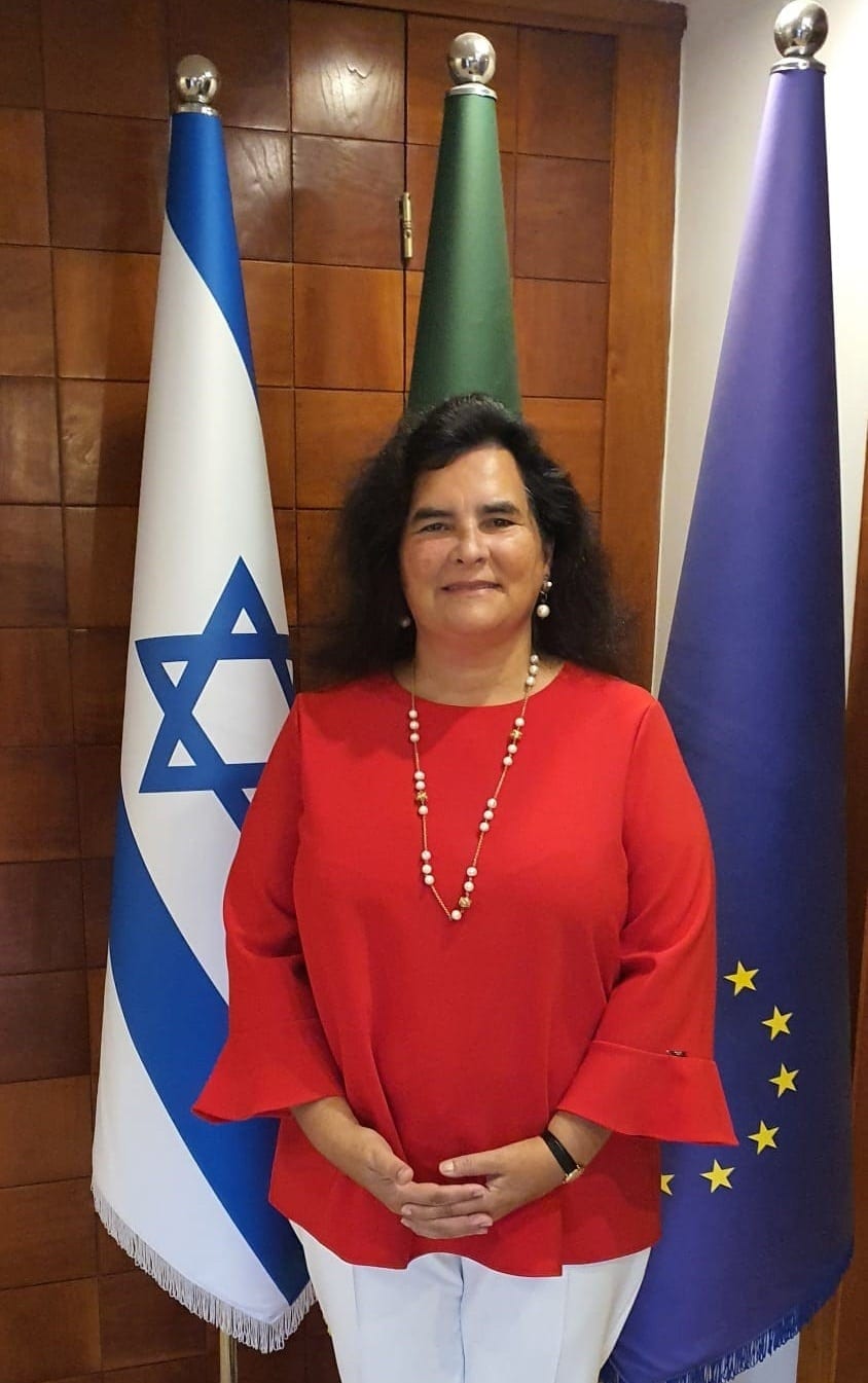 Helena Paiva, Portugal's ambassador to Israel, standing in a red blouse between the flags of Israel, Portugal and the European Union