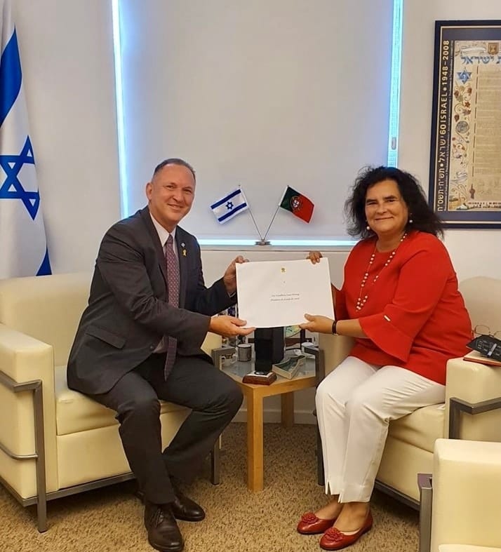 Ambassador Helena Paiva seated with Gil Haskel, holding a document together, beside the flags of Israel and Portugal