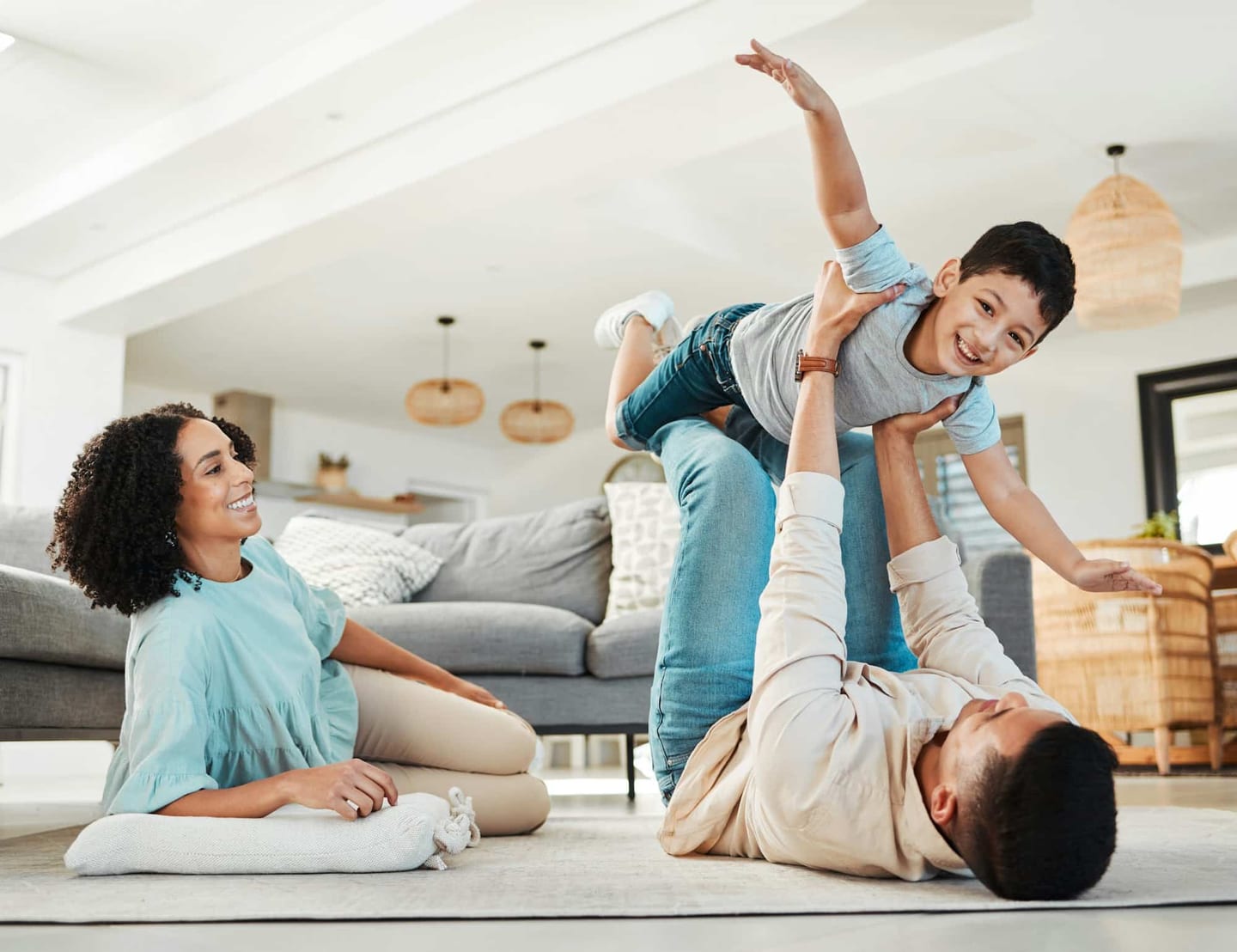 A mother and father playing with their child on the living-room floor — the father lying down lifting the child in the air, the mother sitting beside them smiling