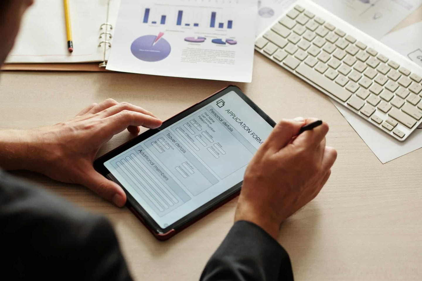 A hand holding a pen and filling in an application form on a tablet, beside a keyboard and documents with charts