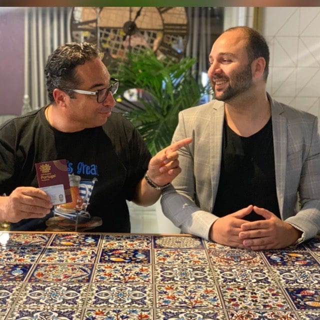 Moti of Easy Nationality (left, in glasses and a black shirt) holding a Portuguese citizen card and pointing at a smiling client in a grey jacket, at a table covered with Portuguese azulejo tiles