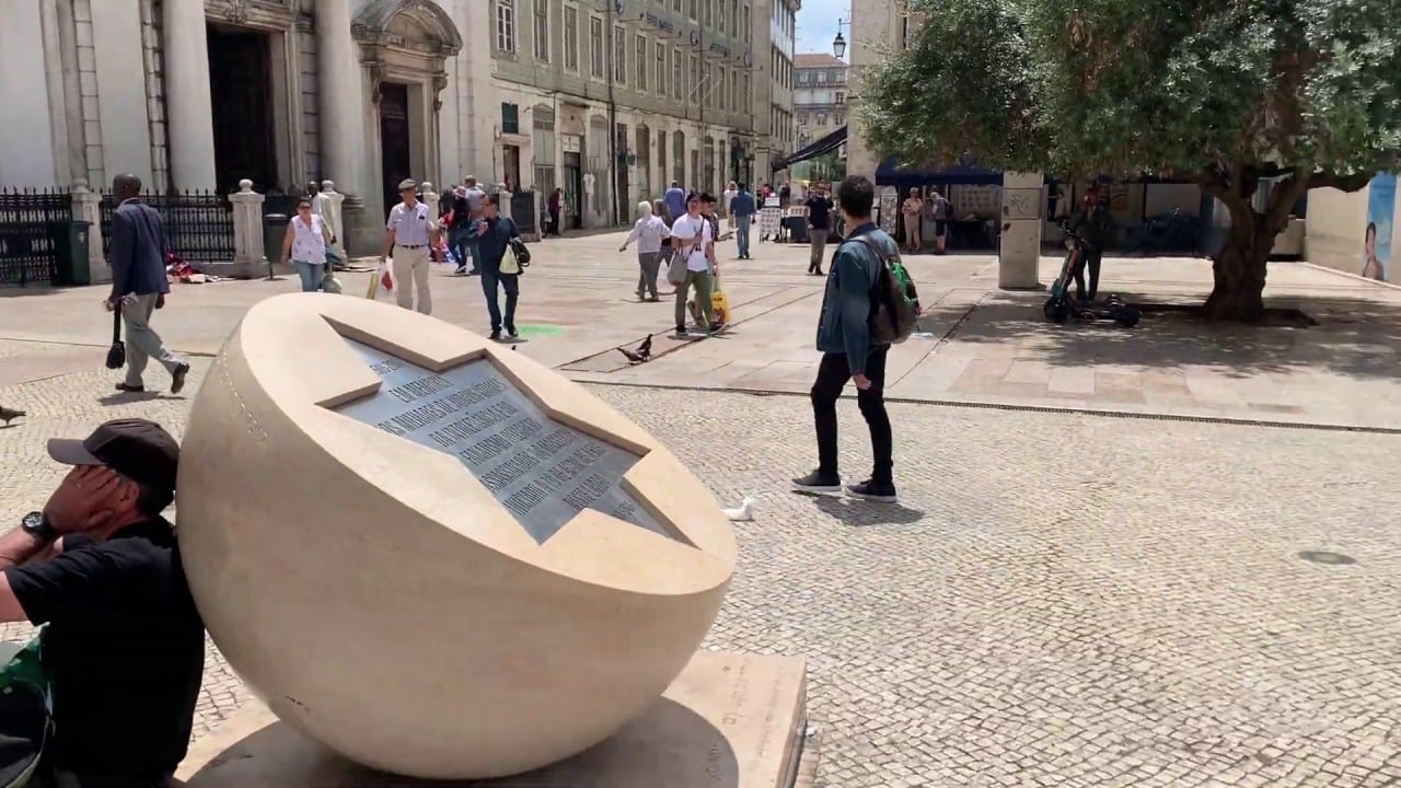 Still from a Portugo tour video — a marble sculpture in a square in Lisbon with visitors around it